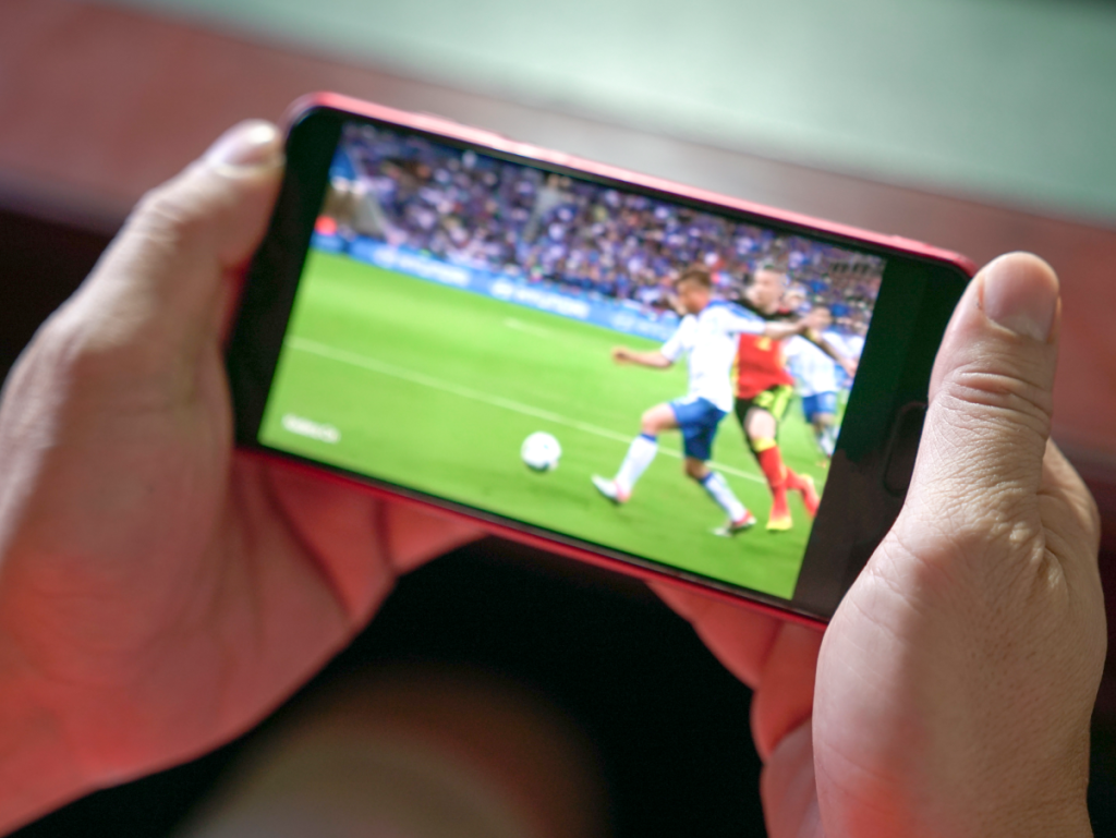 Mão segurando celular em modo paisagem, na tela está passando um jogo de futebol ao vivo.