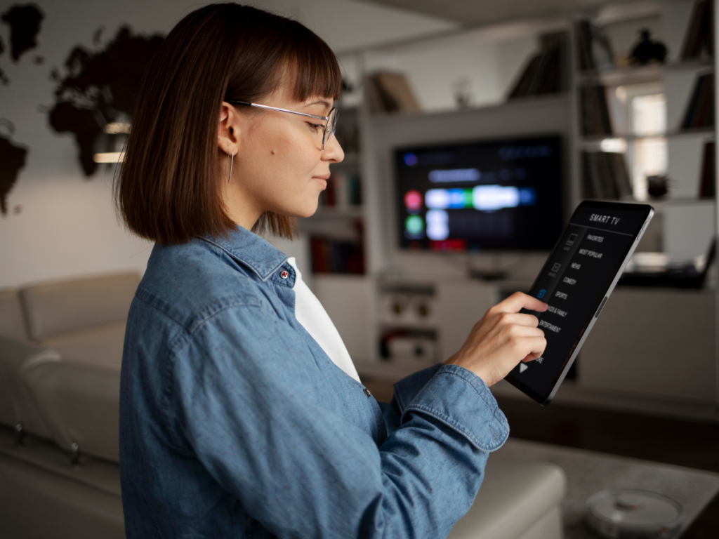 Mulher segurando um tablet e navegando por um menu de canais de IPTV em um ambiente doméstico moderno, com uma Smart TV ao fundo exibindo uma interface de streaming. A imagem representa a experiência de testar um serviço de IPTV antes de contratá-lo.
