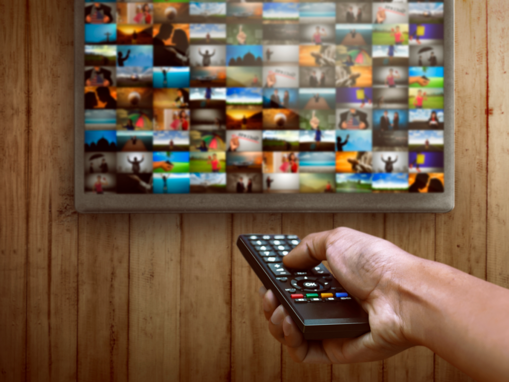 Pessoa segurando um controle remoto apontado para uma Smart TV com uma grade de miniaturas de programas variados na tela, representando a experiência de testar um serviço de IPTV antes de contratar.