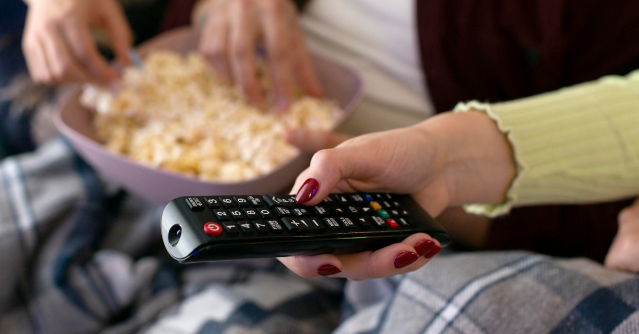 Mão segurando controle remoto enquanto pessoas compartilham uma tigela de pipoca, representando o início de uma maratona de séries em ambiente aconchegante.