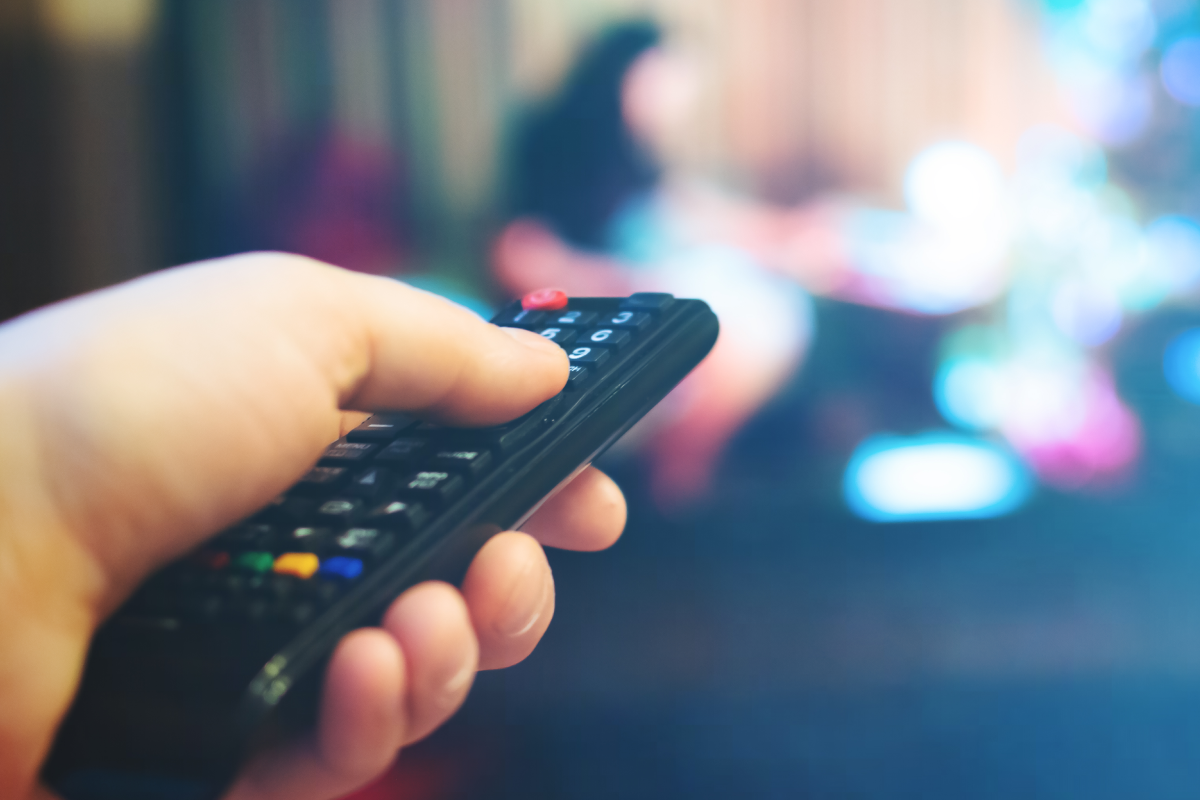 Uma mão segurando o controle remoto de uma TV. Ao fundo, uma televisão ligada, representando a TV Globo ao vivo.