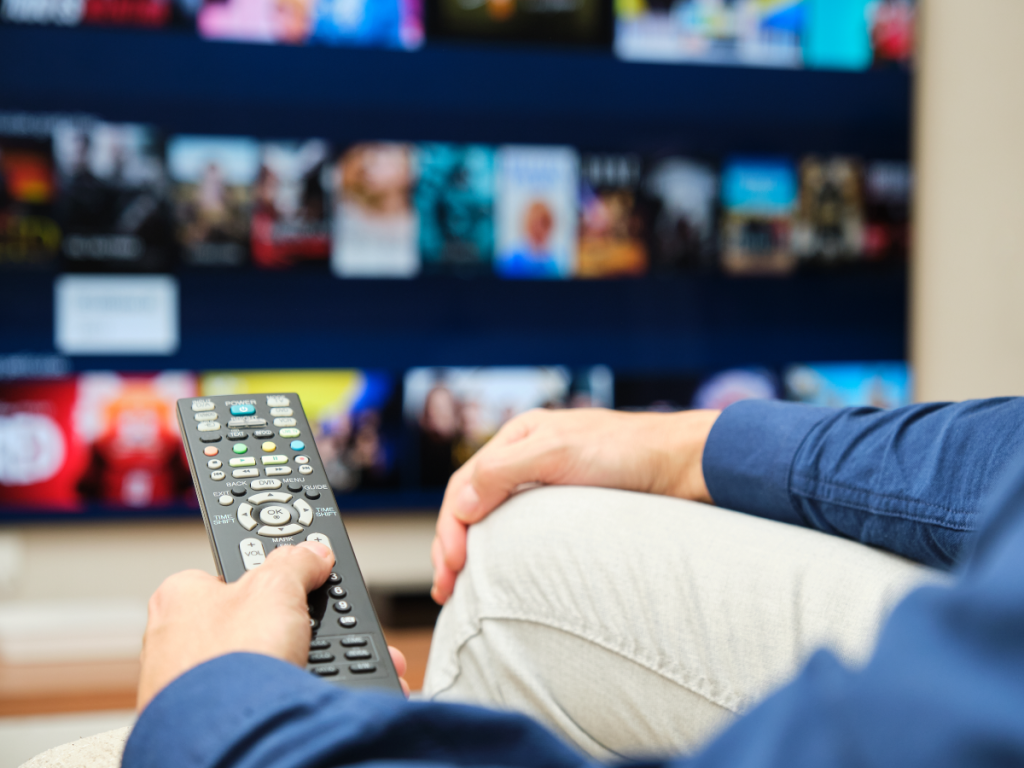 Uma pessoa com controle de TV na mão, com uma TV ao fundo, com um vasto catálogo de canais.