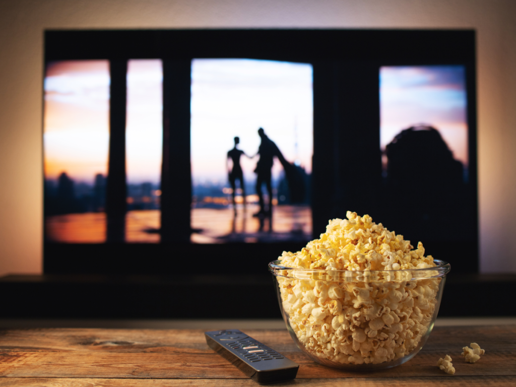 Tigela de pipoca em uma mesa de madeira com controle remoto ao lado, em frente a uma TV exibindo uma cena de filme ao entardecer, simbolizando uma sessão de filmes clássicos em casa.