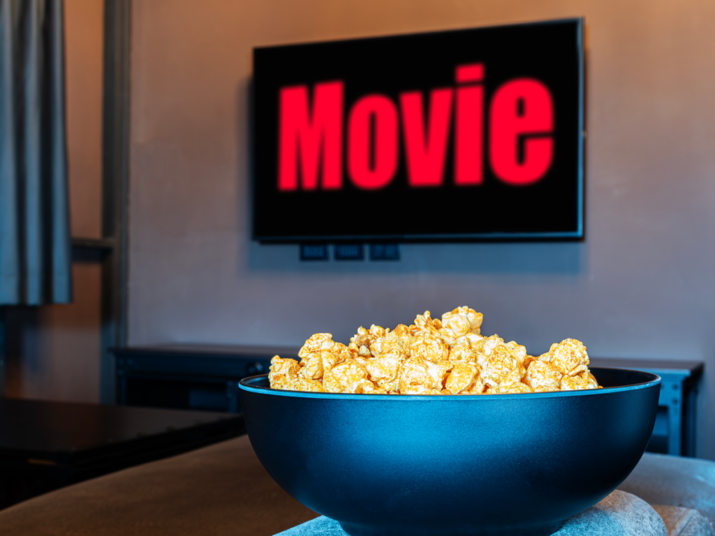 Tigela de pipoca em destaque com uma televisão ao fundo exibindo um filme, simbolizando o conforto de assistir filmes de lançamentos em casa com qualidade de cinema.