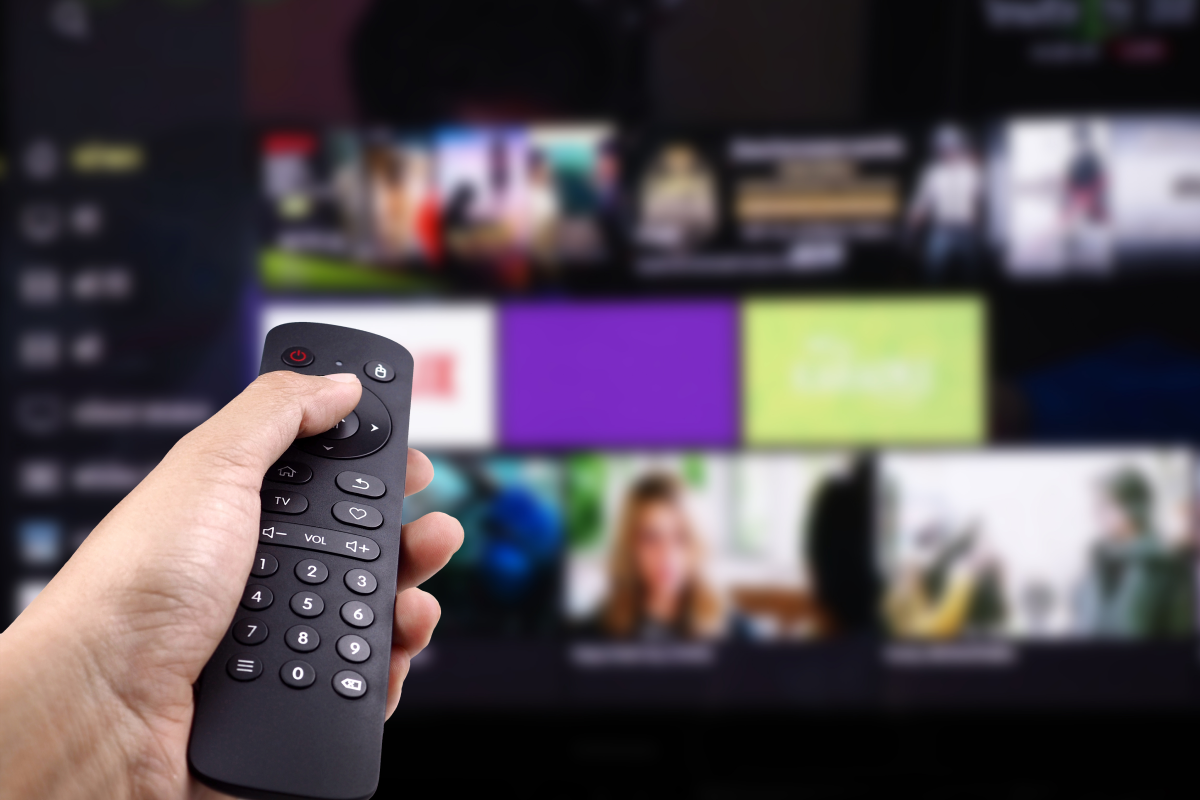 Mão segurando um controle remoto em frente a uma Smart TV com a interface de streaming aberta, representando a navegação por séries online grátis.