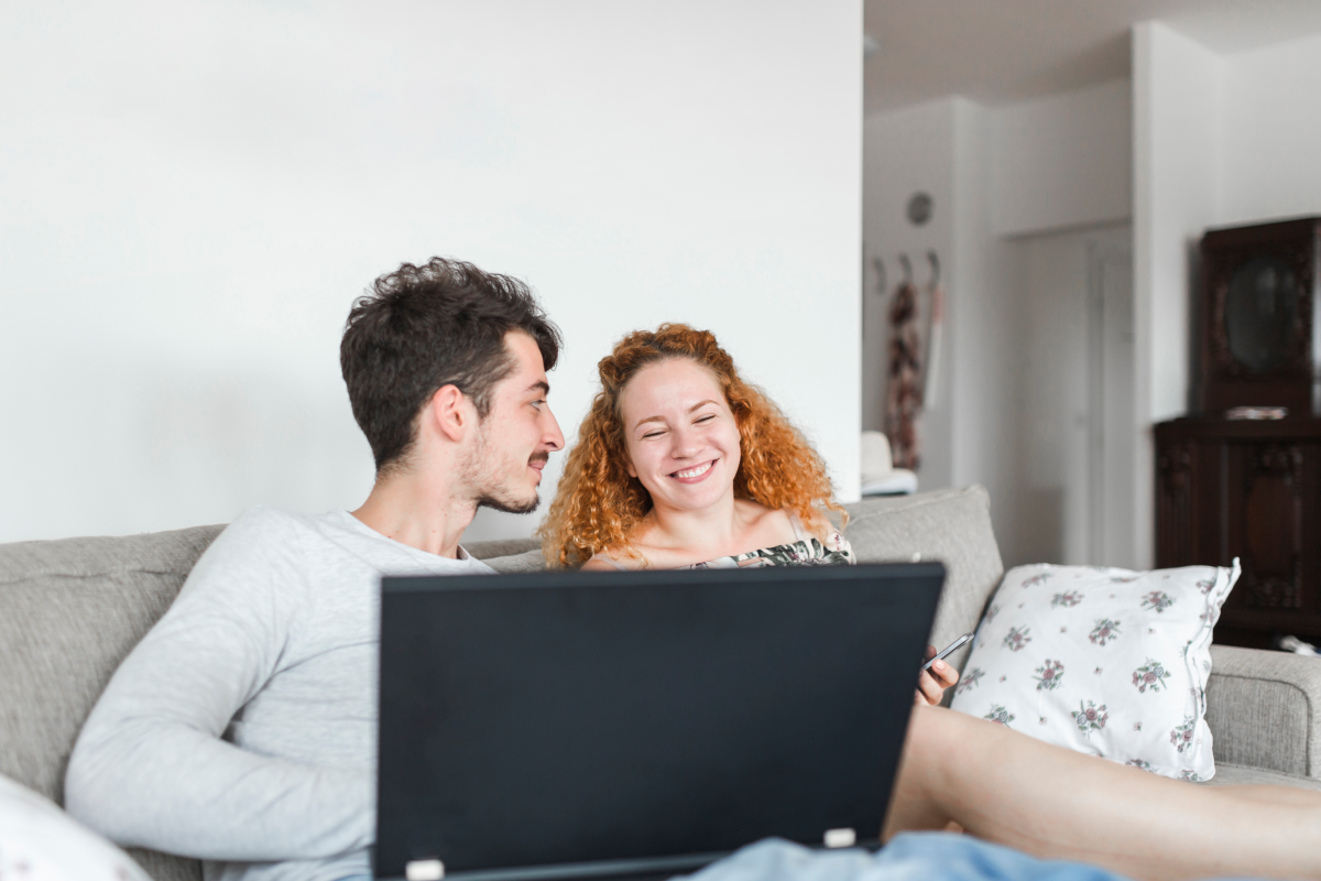 Casal sorridente sentado no sofá assistindo TV online em um notebook, em um ambiente doméstico aconchegante.