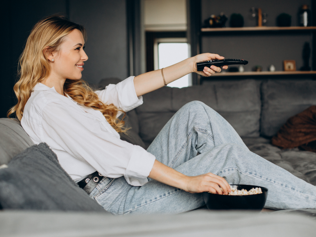 Mulher relaxada no sofá usando controle remoto enquanto assiste TV, representando a praticidade do IPTV instalado diretamente na televisão.