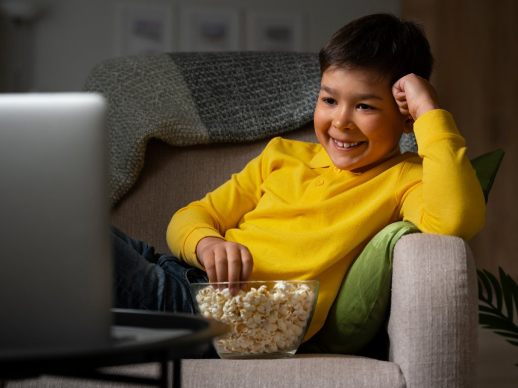 Menino comendo pipoca enquanto assiste canais infantis no IPTV, usando um notebook, sentado confortavelmente em uma poltrona.