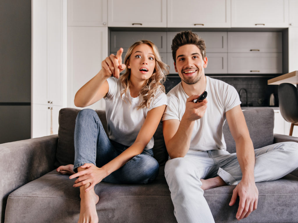 Casal jovem sentado no sofá da sala assistindo televisão juntos, sorrindo e interagindo com o conteúdo exibido na TV, representando o uso do teste IPTV, em um ambiente moderno e iluminado.