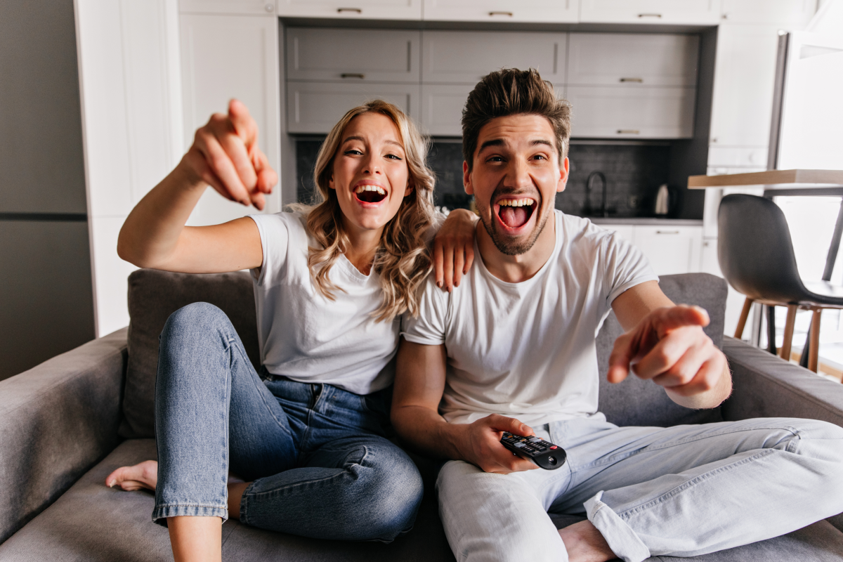 Casal jovem sentado no sofá, sorrindo e apontando para a televisão enquanto assistem a filmes de comédia. Ambos estão vestidos com roupas casuais e demonstram alegria e descontração em um ambiente acolhedor de sala de estar.