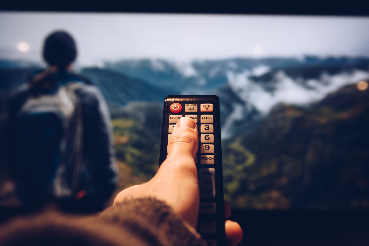 Pessoa segurando um controle remoto apontado para uma TV que exibe uma cena de aventura em alta definição, com paisagens montanhosas e um explorador de mochila ao fundo. A imagem simboliza a experiência de assistir filmes de aventura em casa, com qualidade de cinema.