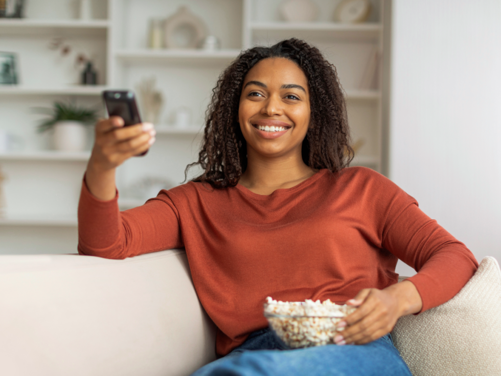 Mulher sorridente assistindo TV em casa com controle remoto e tigela de pipoca, representando o conforto de consumir filmes online.