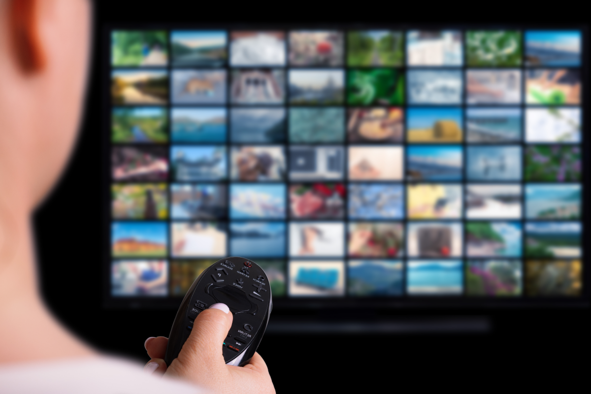 Pessoa segurando um controle remoto enquanto assiste a uma TV com múltiplas miniaturas de filmes e séries, representando o conceito de assistir filmes online grátis via streaming ou IPTV.