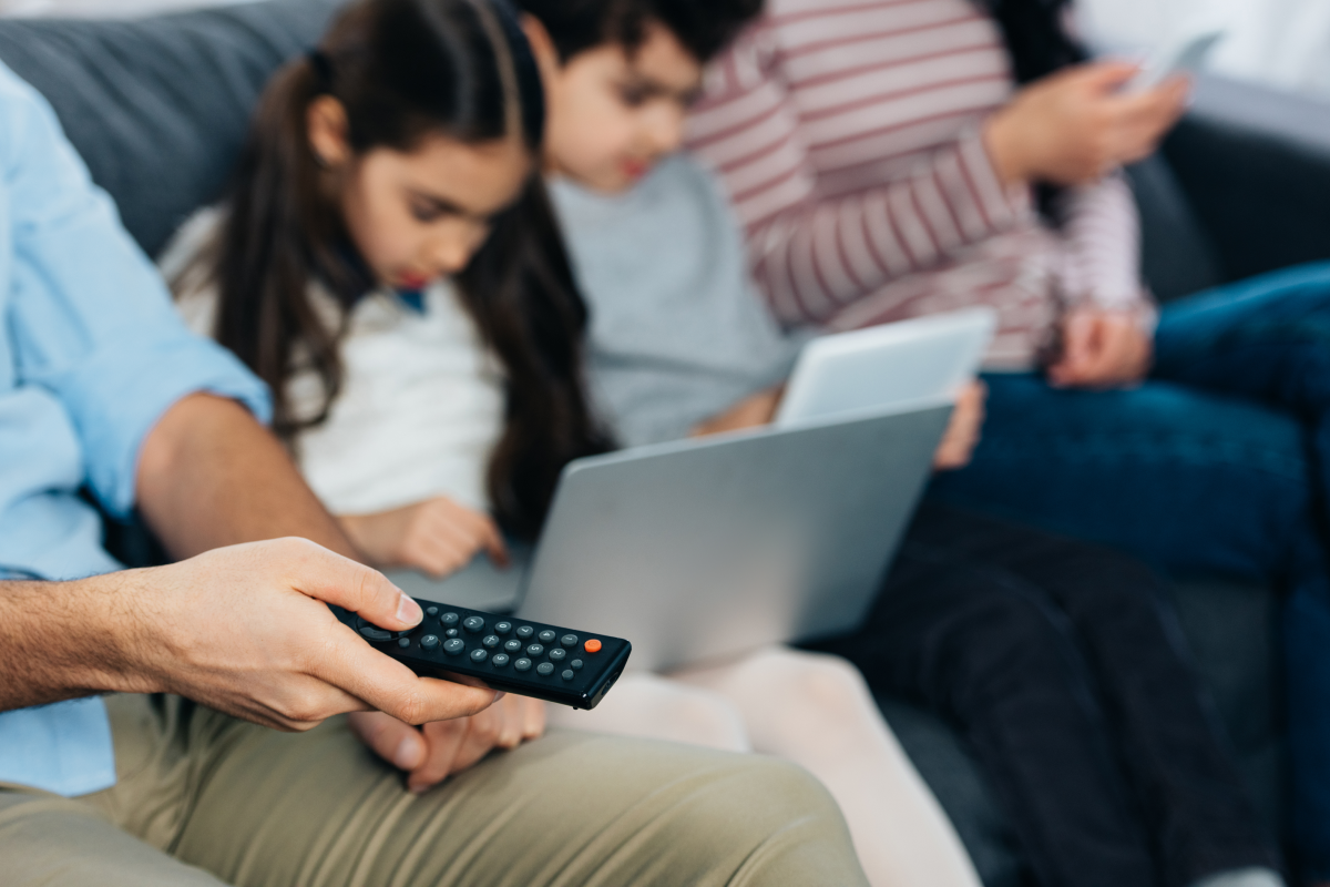 Família reunida no sofá usando controle remoto, notebook e tablet, ilustrando diferentes formas de assistir filmes online gratuitos e pagos.