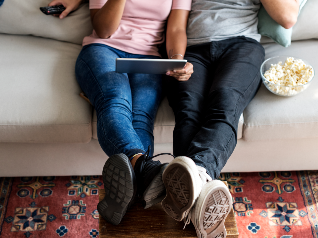 Casal assistindo filmes dublados em casa, usando tablet e compartilhando pipoca no sofá.