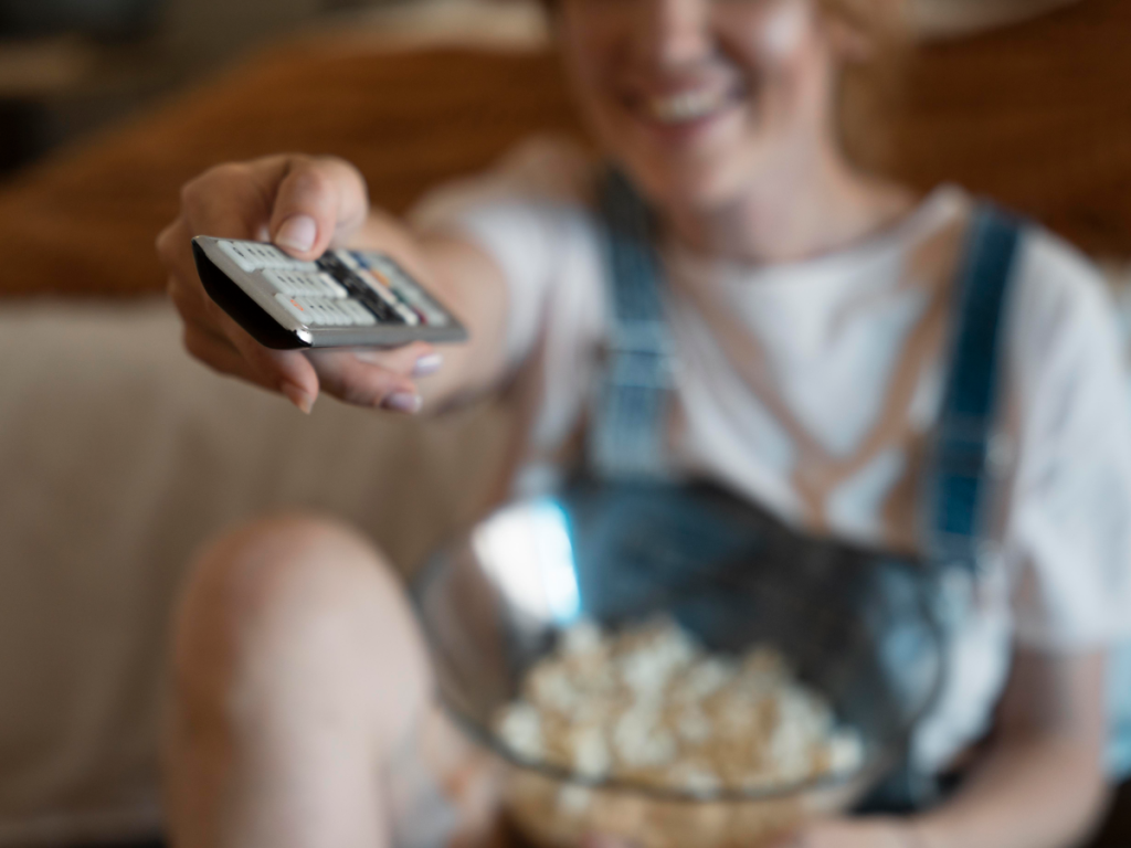 Mulher sorridente em casa segurando um controle remoto em direção à câmera, com um balde de pipoca no colo, simbolizando momentos de lazer assistindo filmes ou T