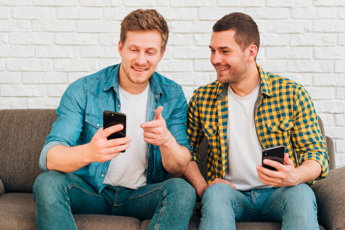 Dois homens sorrindo enquanto usam seus smartphones sentados em um sofá, simbolizando o uso de IPTV no celular.