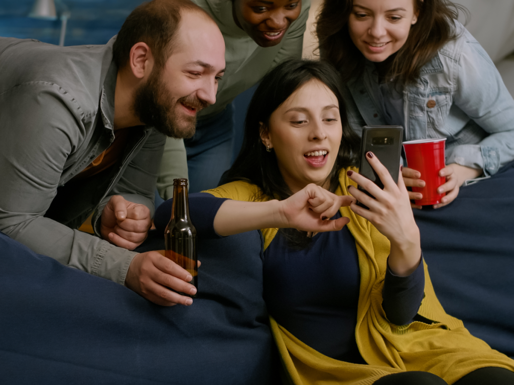 Grupo de amigos reunidos em casa assistindo a conteúdo de TV online em um smartphone, sorrindo e interagindo animadamente com bebidas na mão.