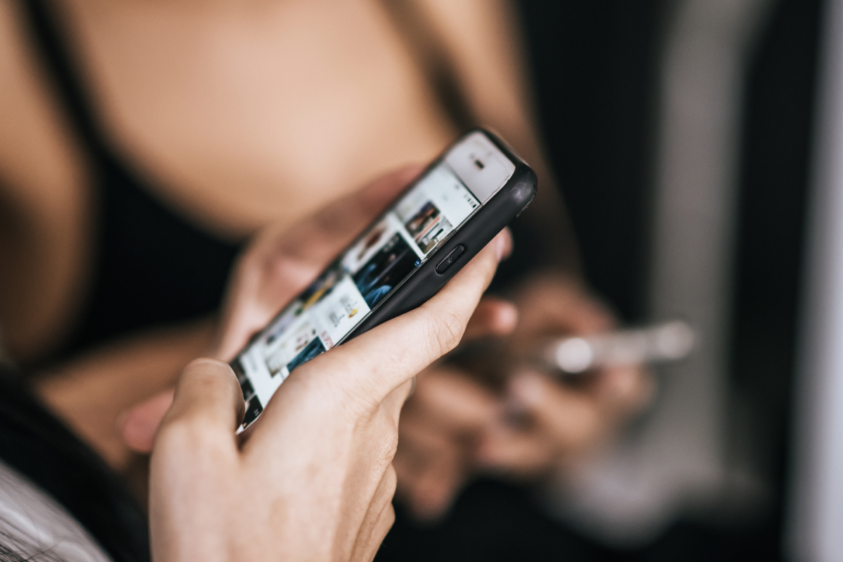 Pessoa segurando um smartphone enquanto navega por uma interface de mídia IPTV, com outra pessoa desfocada ao fundo também usando IPTV no smartphone.
