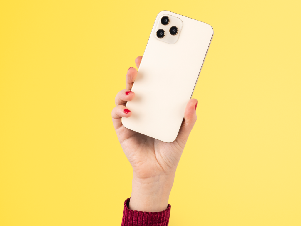 Mão feminina segurando um iPhone branco com três câmeras, em destaque sobre fundo amarelo. Conceito de mobilidade e tecnologia.