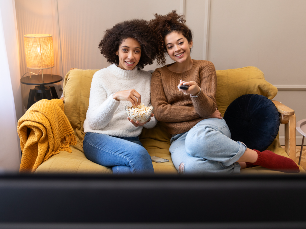 Duas mulheres relaxam no sofá assistindo a conteúdos via IPTV com pipoca na mão e controle remoto, simbolizando o entretenimento digital em 2025