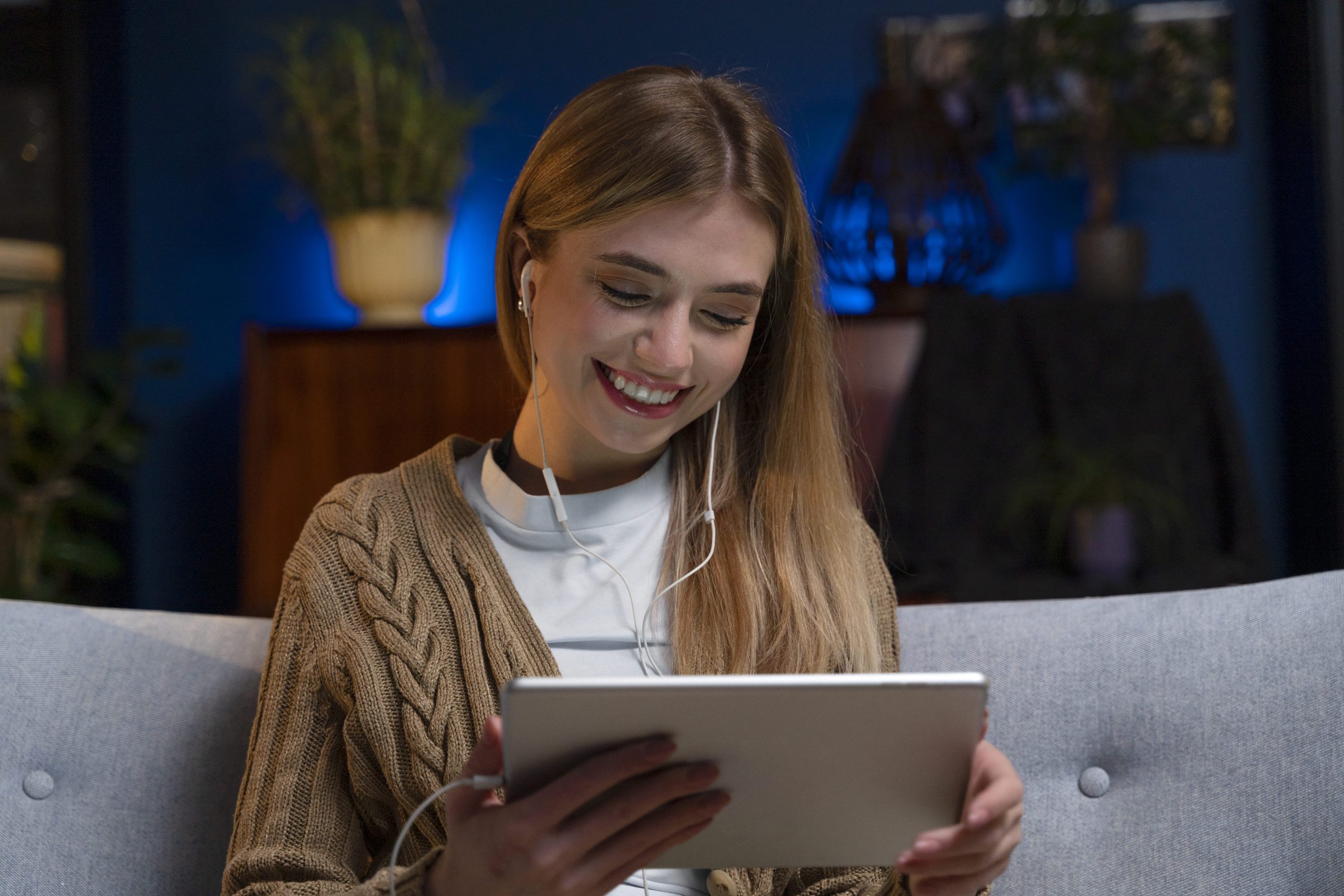 Mulher sorridente sentada no sofá, assistindo conteúdo IPTV no tablet com fones de ouvido, em ambiente aconchegante e iluminado.