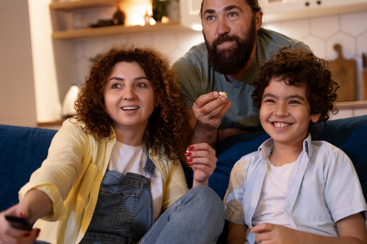 Família sorridente assistindo canais infantis no IPTV junta no sofá, comendo pipoca e se divertindo em casa.
