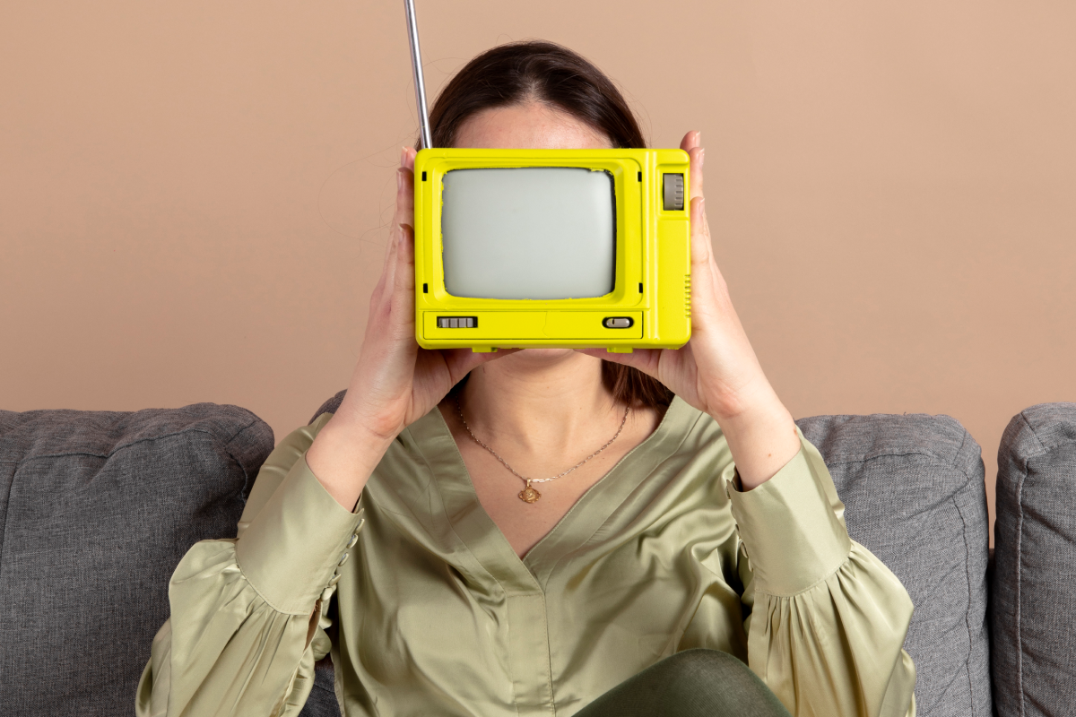 Mulher sentada em um sofá segurando uma pequena televisão amarela retrô em frente ao rosto, simbolizando a nostalgia da IPTV e TV por assinatura tradicional em contraste com as novas formas de assistir conteúdo com o IPTV.