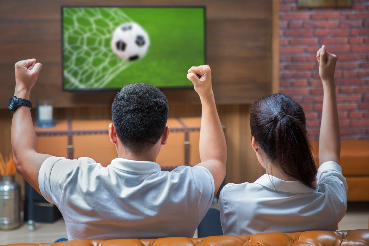 Casal comemorando um gol enquanto assiste a uma partida de futebol ao vivo na TV, em um ambiente aconchegante de sala de estar. A imagem representa a emoção de acompanhar jogos em alta qualidade através do IPTV.