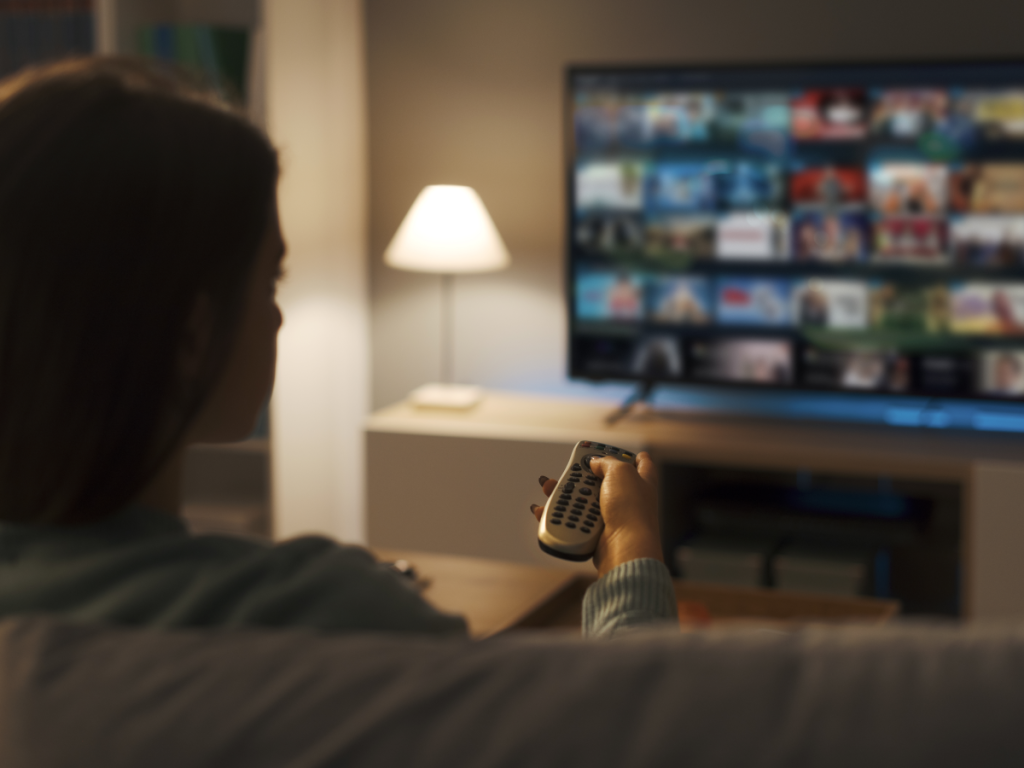 Mulher assistindo serviços de streaming na TV com controle remoto em mãos, representando a variedade de plataformas disponíveis em 2025.