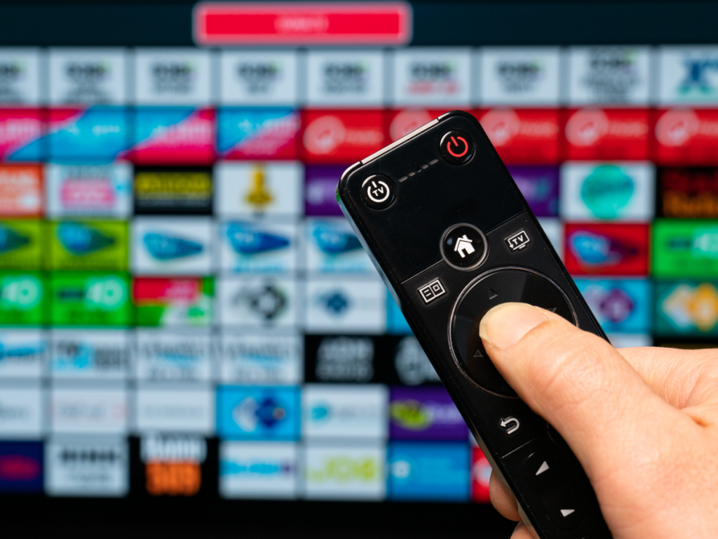 Mão segurando controle remoto com interface de IPTV ao fundo, destacando a variedade de canais disponíveis via IPTV.