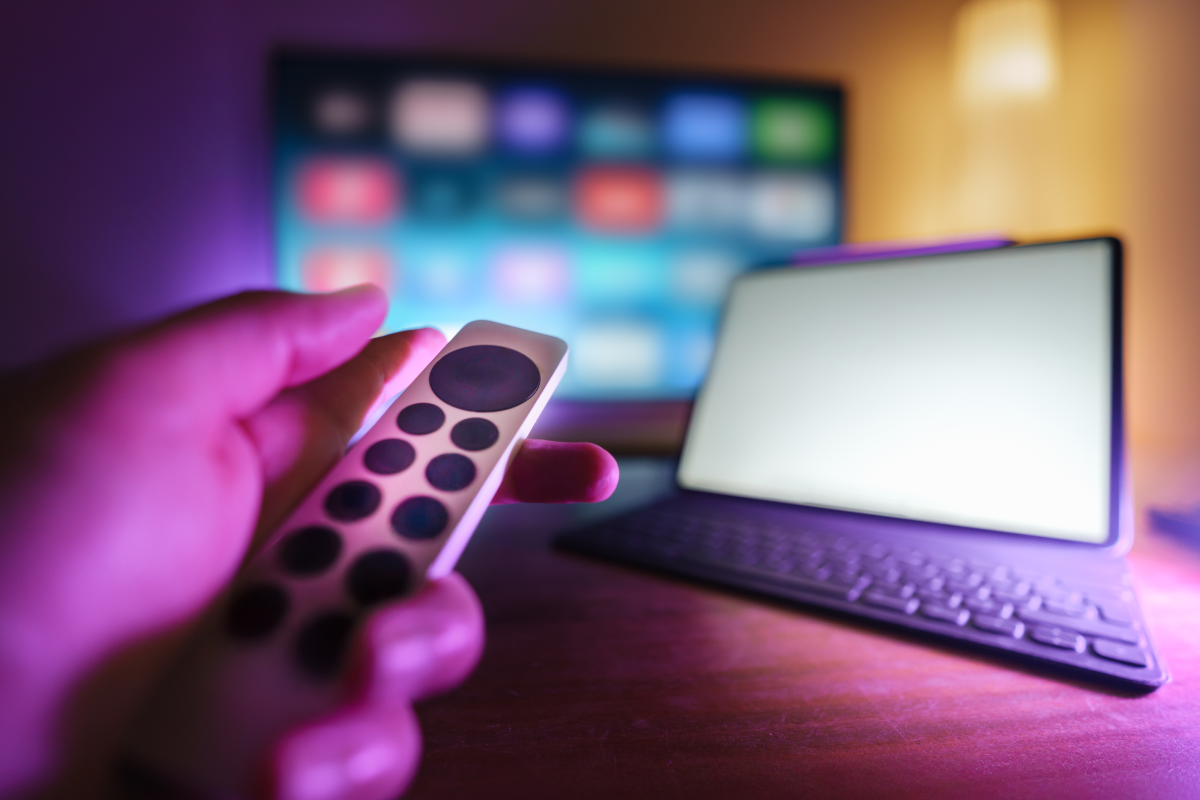 Pessoa segurando um controle remoto diante de uma TV com interface de aplicativos coloridos desfocada, ao lado de um tablet sobre a mesa, simbolizando acesso a conteúdos por IPTV em diferentes dispositivos durante o teste grátis IPTV.