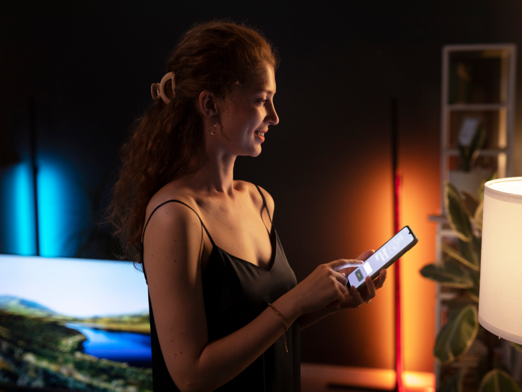 Mulher usando o celular em casa, com a TV ligada ao fundo, representando o uso do IPTV no celular para entretenimento doméstico.
