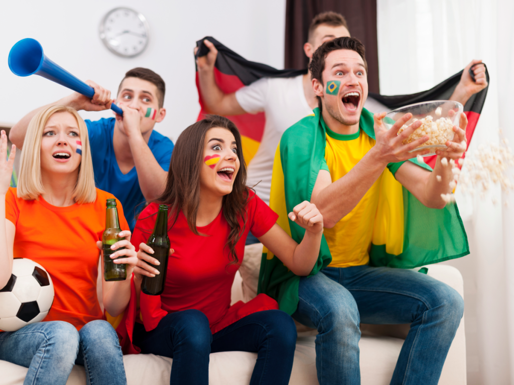 Grupo de amigos assistindo futebol ao vivo via IPTV, vibrando durante partida com bandeiras e pinturas faciais dos times.