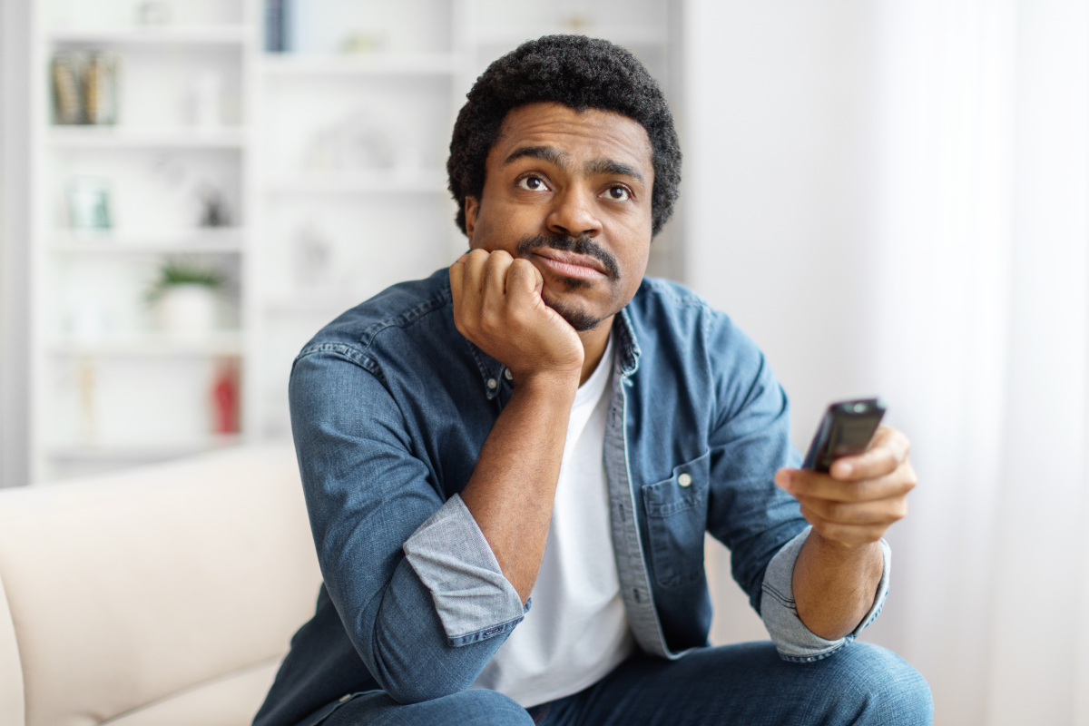 Homem pensativo sentado no sofá segurando controle remoto, representando dúvida sobre qual serviço de IPTV confiável contratar.