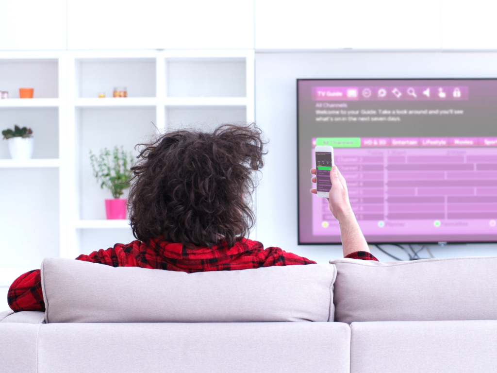 Uma pessoa com celular na mão e ao fundo uma TV, representando o uso de lista grátis IPTV em ambos os dispositivos.