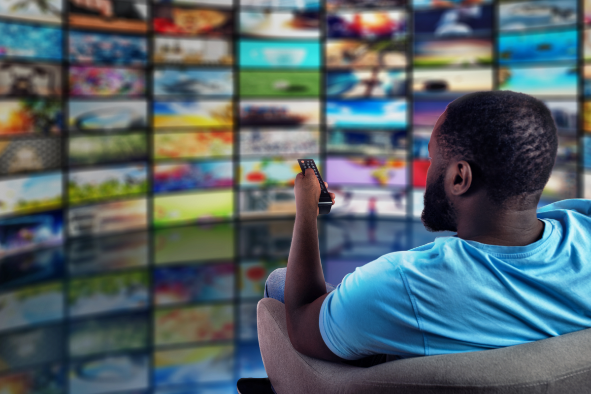 Homem assistindo a uma parede de canais de TV em uma interface digital de IPTV, usando controle remoto, representando a variedade de conteúdos disponíveis em aplicativos IPTV.
