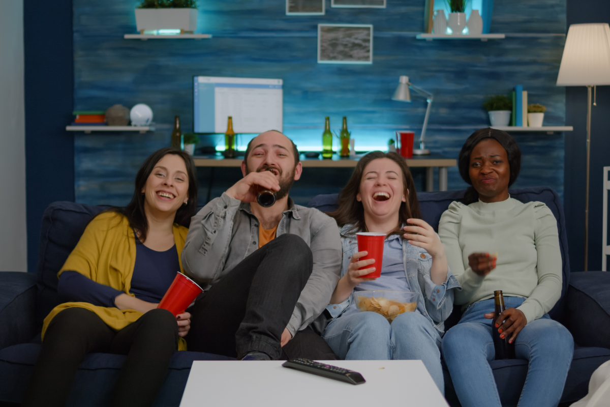 Grupo de amigos de diferentes etnias assistindo a uma série de comédia juntos no sofá, rindo e se divertindo com petiscos e bebidas – momento descontraído típico de uma maratona de séries em casa.