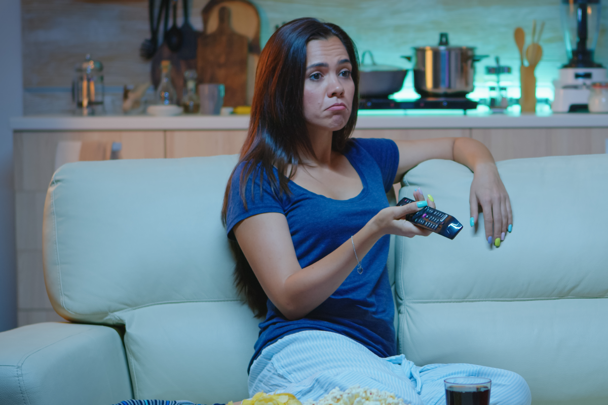 Mulher sentada no sofá com controle remoto, enfrentando travamentos enquanto tenta assistir TV, representando frustração comum com pacotes IPTV instáveis.
