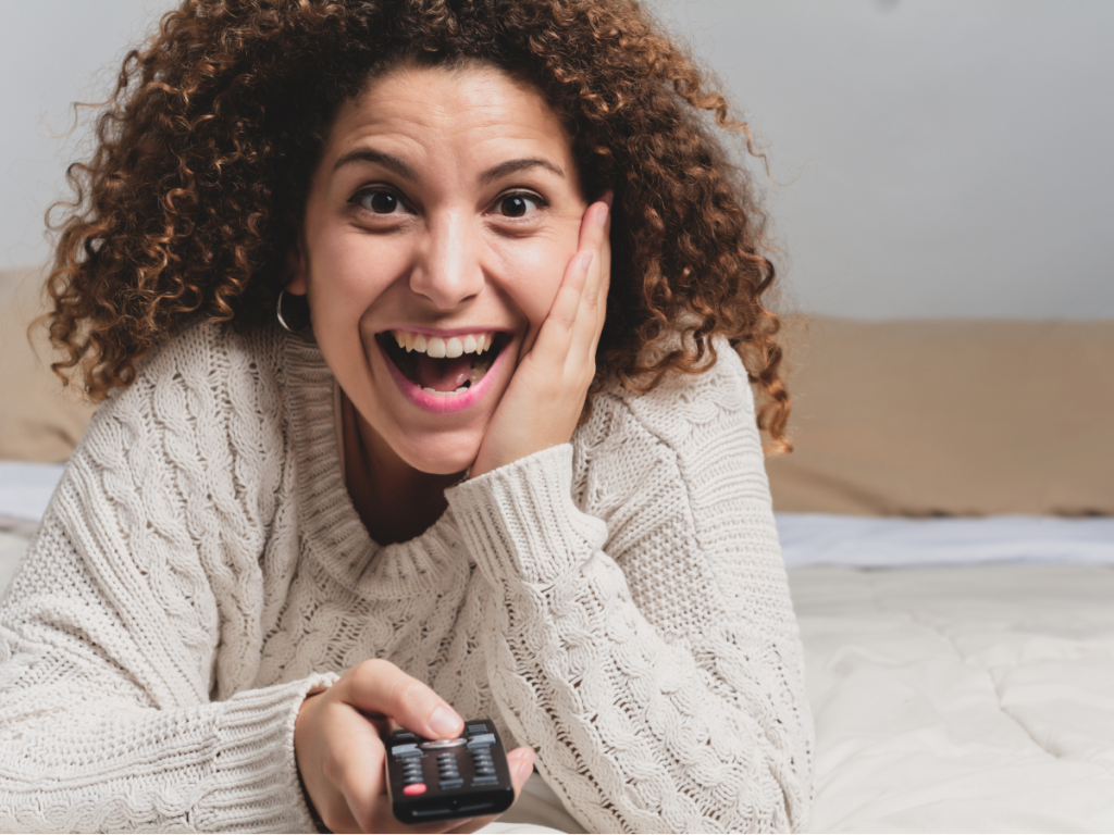 Mulher sorridente deitada na cama com controle remoto na mão, assistindo a uma série de comédia e se divertindo.
